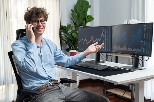 Profile of smiling young stock trader with curly hair looking at camera while calling to trader against financial exchange screen at office. Concept of dynamic business digital investment. Gusher.