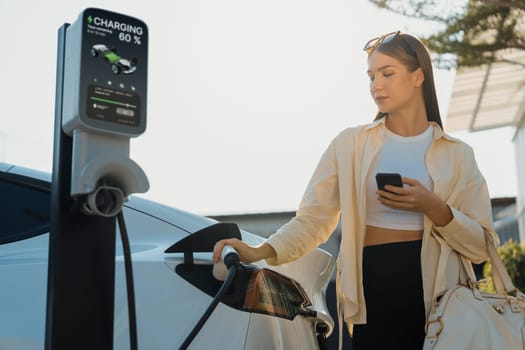 Young woman holding shopping bag and use smartphone to pay for electricity for recharging EV car battery from charging station at city mall parking lot. Modern woman go shopping by eco car. Expedient