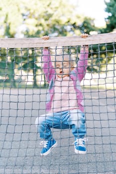 Little girl hangs on the edge of a tennis net with her legs folded. High quality photo