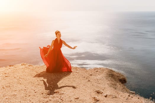 Side view a Young beautiful sensual woman in a red long dress posing on a rock high above the sea during sunrise. Girl on the nature on blue sky background. Fashion photo.