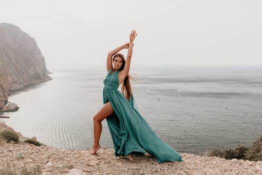 Side view a Young beautiful sensual woman in a mint long dress posing on a volcanic rock high above the sea during sunset. Girl on the nature on overcast sky background. Fashion photo