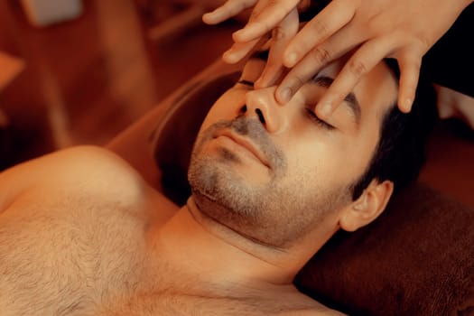 Caucasian man enjoying relaxing anti-stress head massage and pampering facial beauty skin recreation leisure in warm candle lighting ambient salon spa in luxury resort or hotel. Quiescent