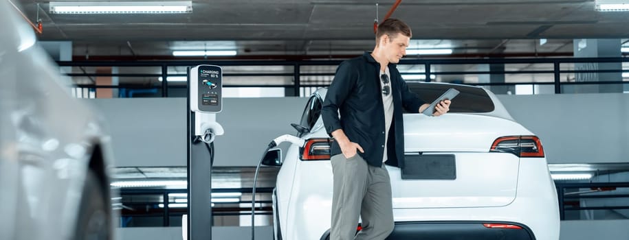 Young man travel with EV electric car to shopping center parking lot charging in downtown city showing urban sustainability lifestyle by green clean rechargeable energy of electric vehicle innards