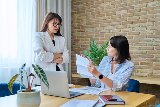 Two serious business women talking working in modern office, young female reading contract document, communicating colleague. Business, ceo, work, law, finance, mentoring, consulting, teamwork, job concept