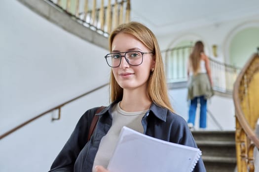 Portrait of young teenage girl student posing inside an educational building, standing on stairs. Smiling female with bag, textbook looking at camera. Youth, education, college, university