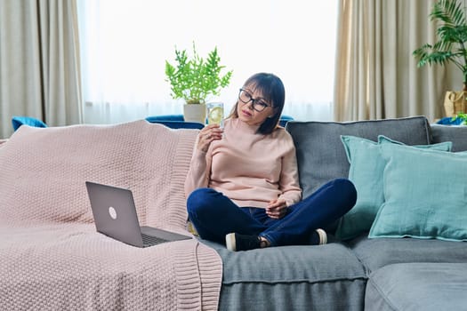 Middle aged 40s relaxed woman at home on sofa in living room, with glass of water with lemon, with laptop. Relaxation, leisure, lifestyle, food vitamins for autumn winter season