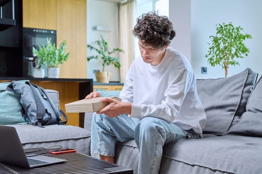 Young male unpacking an online purchase, sitting on couch at home. Internet online shopping, delivery, youth concept