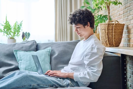 Young guy 18-20 years old resting at home, sitting on sofa in living room, typing laptop. Internet online technology, study leisure communication concept