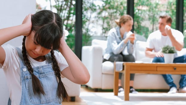 Stressed and unhappy young girl huddle in corner, cover her ears blocking sound of her parent arguing in background. Domestic violence at home and traumatic childhood develop to depression. Synchronos