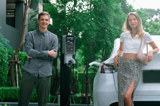 Young couple travel with EV electric car charging in green sustainable city outdoor garden in summer shows urban sustainability lifestyle by green clean rechargeable energy of electric vehicle innards