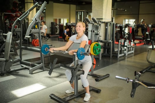 Woman training biceps with a barbell in the gym. Strength training with heavy equipment.
