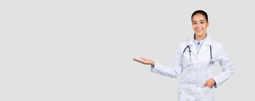 Cheerful european millennial doctor in white coat, with stethoscope hold copy space on hand, isolated on gray studio background, panorama. Medical ad and offer, health care presentation, sale