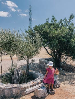 Little girl with a basket stands near a stone flower bed and looks at a tree. High quality photo