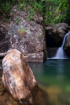 Long exposure photo of a hidden waterfall in a rocky riverbed within a tropical rainforest, featuring blurred rocks upfront.