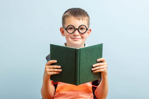 Little schoolboy in eyeglasses is reading a book on blue background. Education and school concept.