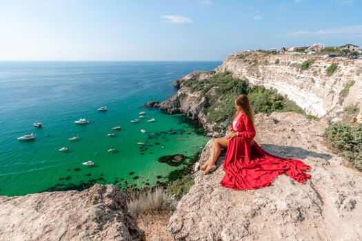 Woman red dress sea. Happy woman in a red dress and white bikini sitting on a rocky outcrop, gazing out at the sea with boats and yachts in the background