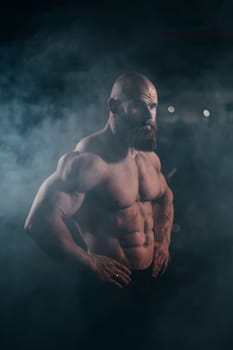 A muscular bald man poses shirtless in the dark amid smoke. Vertical photo