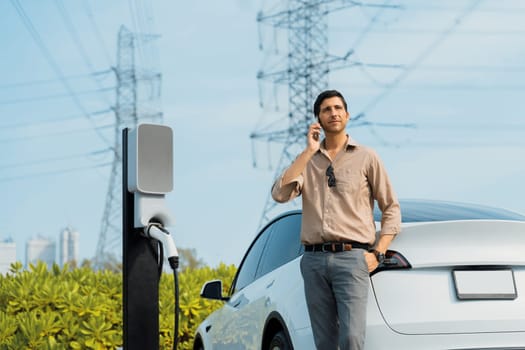 Man talking on the phone while recharge EV car battery at charging station connected to power grid tower electrical as electrical industry for eco friendly car utilization.Expedient