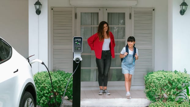 Happy little young girl learn about eco-friendly and energy sustainability as she help her mother recharge electric vehicle from home EV charging station. EV car and modern family. Panorama Synchronos