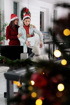 Businesswoman wearing red Christmas hat holding box filled with desk belongings saying goodbye to coworker after being laid off. Upset worker fired from job getting consoled by colleague