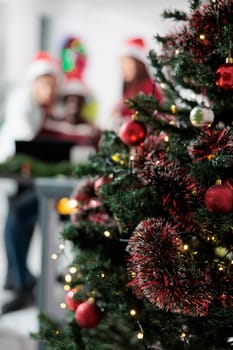 Close up focus shot on office xmas tree in festive decorated workspace with business coworkers working in the background. Christmas ornate company workplace during winter holiday season