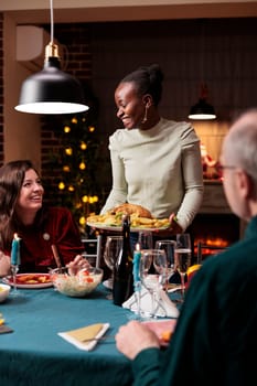 People celebrating christmas eve together with festive dishes and wine, enjoying winter seasonal event with family. Men and women meeting near fireplace to celebrate traditional day with xmas tree.