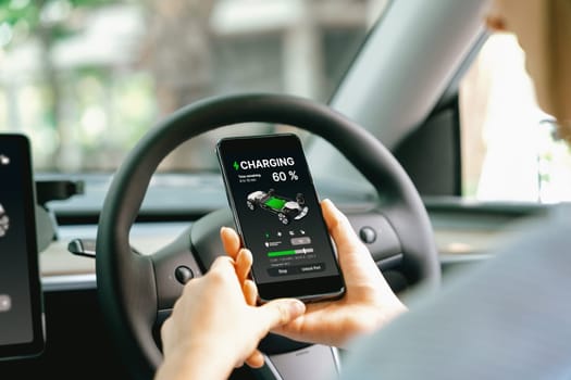 Young woman inside the vehicle, checking EV vehicle's application on battery recharging electricity status display on smartphone screen in modern EV car on her road trip journey. Exalt