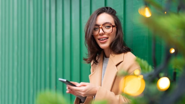 Woman hold smartphone leaning wall looking at camera Christmas light. Portrait happy smiling young adult businesswoman wear glasses and stylish coat standing outdoor. Copy space