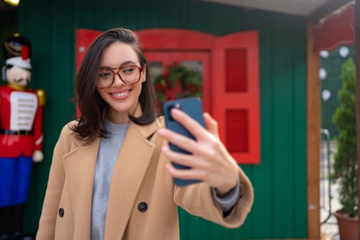 Woman in coat and glasses takes selfies in front of Christmas store. Smiling female taking photo of herself with phone and creating memorie. Woman in coat taking selfie by phone outside