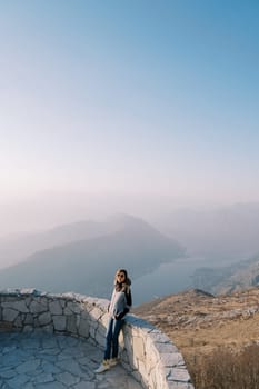 Woman in a jacket stands on an observation deck in the mountains above the bay in the fog. High quality photo