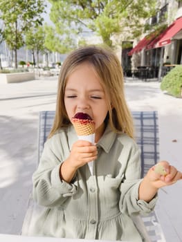 Little girl eating popsicles in a waffle cone while sitting on a chair outdoors. High quality photo
