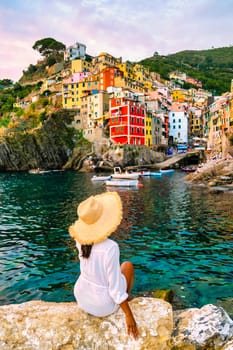 Riomaggiore Cinque Terre Italy, colorful village harbor front by the ocean, young Asian woman with a hat watching the sunset at waterfront looking out over Riomaggiore village Cinque Terre Italy