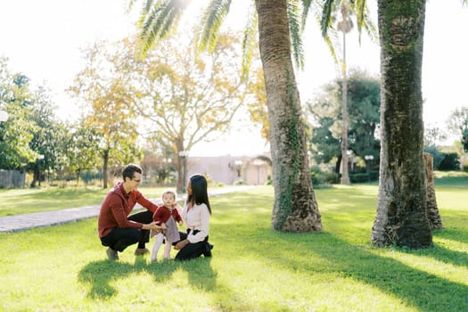 Parents squatting and looking at little girl in sunny park. High quality photo