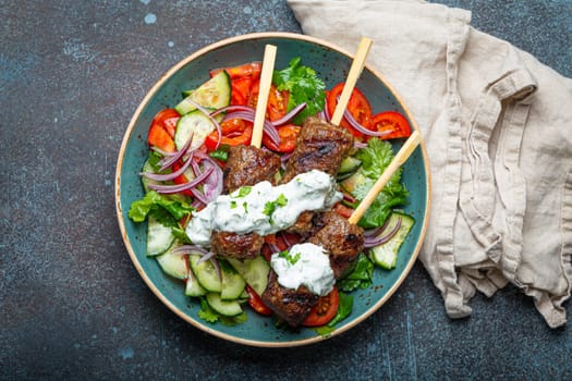 Grilled skewer meat beef kebabs on sticks served with fresh vegetables salad on plate on rustic concrete background from above. Traditional Middle Eastern and Turkish dish Kebab