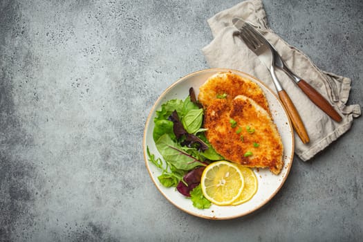 Crispy panko breaded fried chicken fillet with green salad and lemon on plate on gray rustic concrete background table from above. Japanese style deep fried coated chicken breasts, space for text.