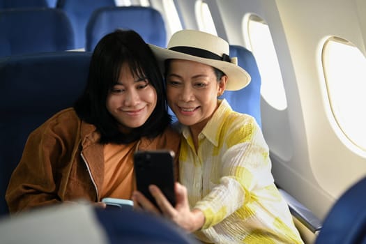 Happy 60s woman and daughter taking photo with smartphone in passenger airplane. Transportation, travel and summer vacation.