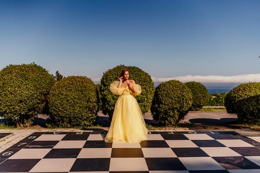 Woman yellow dress chess. A beautiful woman in a long puffy yellow dress poses on a chessboard in the park