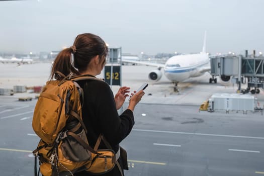 Happy Asian tourist uses mobile smartphone with travel backpack while waiting for flight in airport.