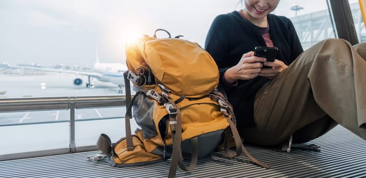 Happy Asian tourist uses mobile smartphone with travel backpack while waiting for flight in airport.