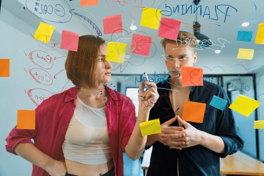 Couple of businesspeople discussing and brainstorming about solving marketing problem in front of glass board at business meeting by using mind map and sticky notes. Working together. Immaculate.