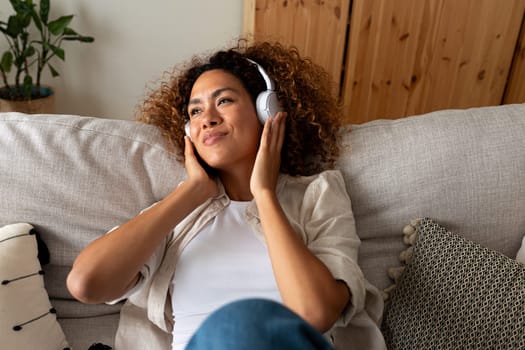 Relaxed young multiracial woman sitting on the sofa listening to music with wireless headphones. Lifestyle concept.