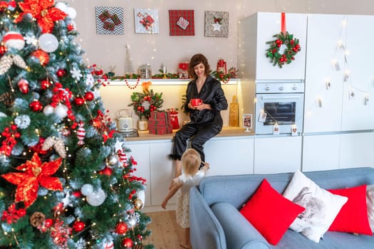 Christmas kitchen mom daughter. They are sitting on a table and drinks from a red mug in a decorated kitchen for christmas