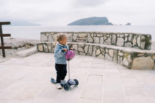 Little girl holds on to the steering wheel of a scooter while standing on the seashore and looks into the distance. High quality photo