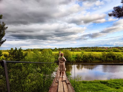 Tourist girl walks along an old wooden suspension bridge over the river and the sky with disturbing clouds. Suicide in nature. The concept of depression and hopelessness of life