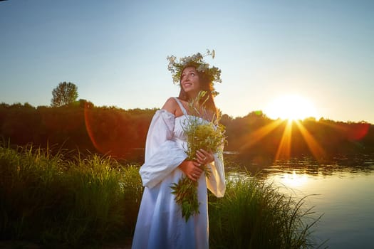 Adult mature brunette woman in a white dress, sundress and a wreath of flowers in summer by the water of river or lake in evening at sunset. Celebration of the Slavic pagan holiday of Ivan Kupala