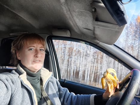 Portrait of female driver in solo journey. Adult mature middle aged woman holding steering wheel and a banana. Eating while driving for to stay awake. Lady girl with food while travel or trip by car