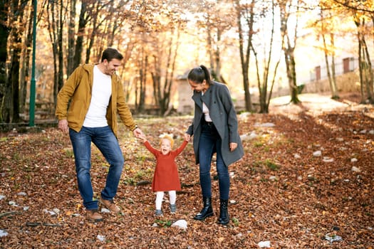 Mom and dad lead by the hands laughing little girl in the autumn park. High quality photo
