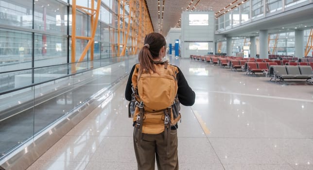 A young Asian woman stands behind in casual clothes and carries a backpack. The schedule can be found on the airport station's scoreboard at weekend..
