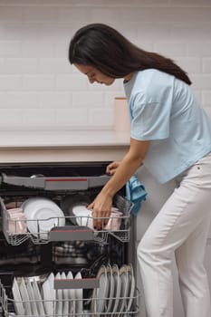 pretty woman loading dishes to open automatic built-in dishwasher machine with clean utensils inside in modern white kitchen.