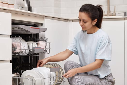 happy woman unloading plate from open automatic built-in dishwasher machine with clean utensils inside in modern white kitchen.